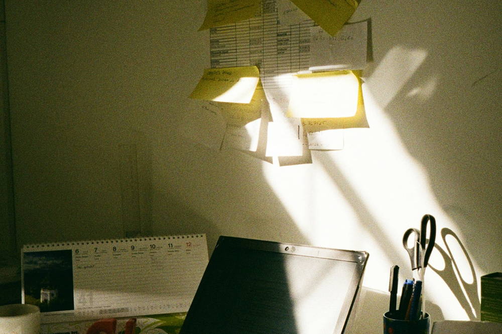 A desk in afternoon light with reminder notes and lists taped across the wall above it